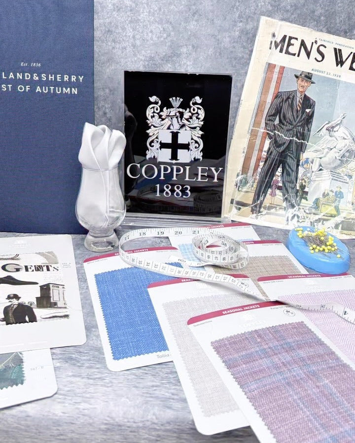 A styled flat-lay of vintage and fashion-related items on a textured gray surface, including a blue book, a black Coppley 1883 card, a magazine page, fabric swatches, a small white figurine, and a blue pin cushion.