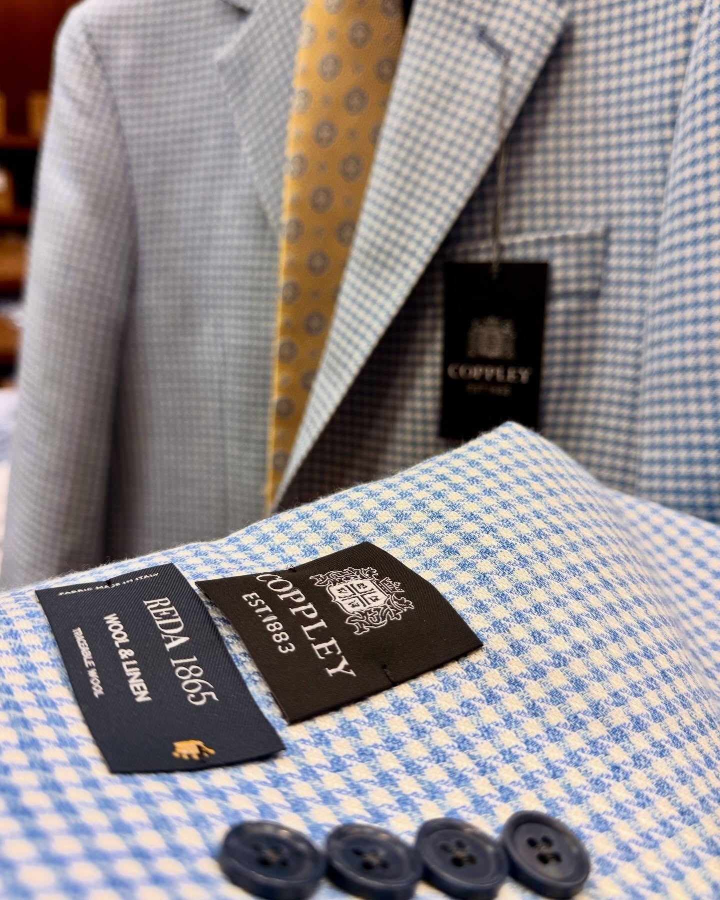 Close-up of a Copley suit jacket in a light blue and white gingham pattern, styled with a yellow patterned tie. The jacket features a black Copley brand label and a matching black pocket tag, with four dark buttons in the foreground.