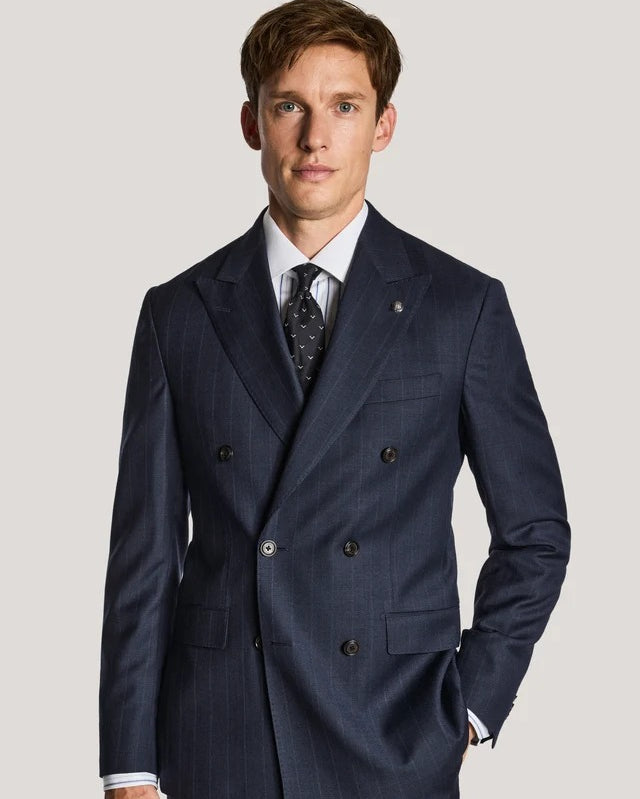 A man wearing a navy blue double-breasted suit with subtle pinstripes, a white dress shirt, and a patterned tie, standing against a light gray studio background.