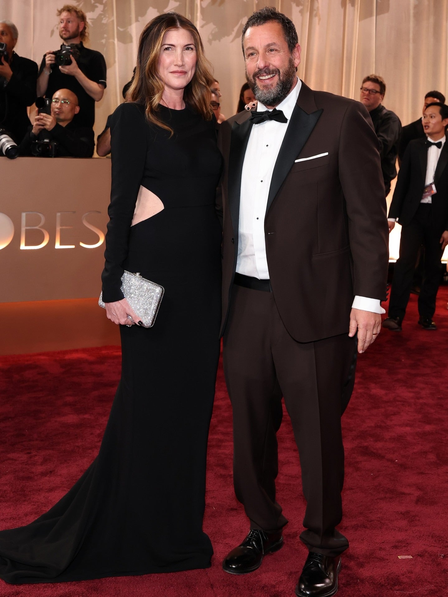 A man and a woman posing together on a red carpet at an awards show. The woman is wearing a floor-length black gown with side cutouts and holding a silver clutch, and the man is wearing a dark brown tuxedo with a white shirt and black bow tie.