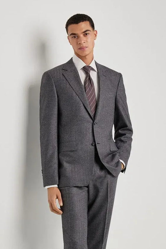 A man modeling a grey two-piece suit with a subtle micro-check pattern, paired with a white dress shirt, a striped tie, and black dress shoes, standing against a light grey studio background.