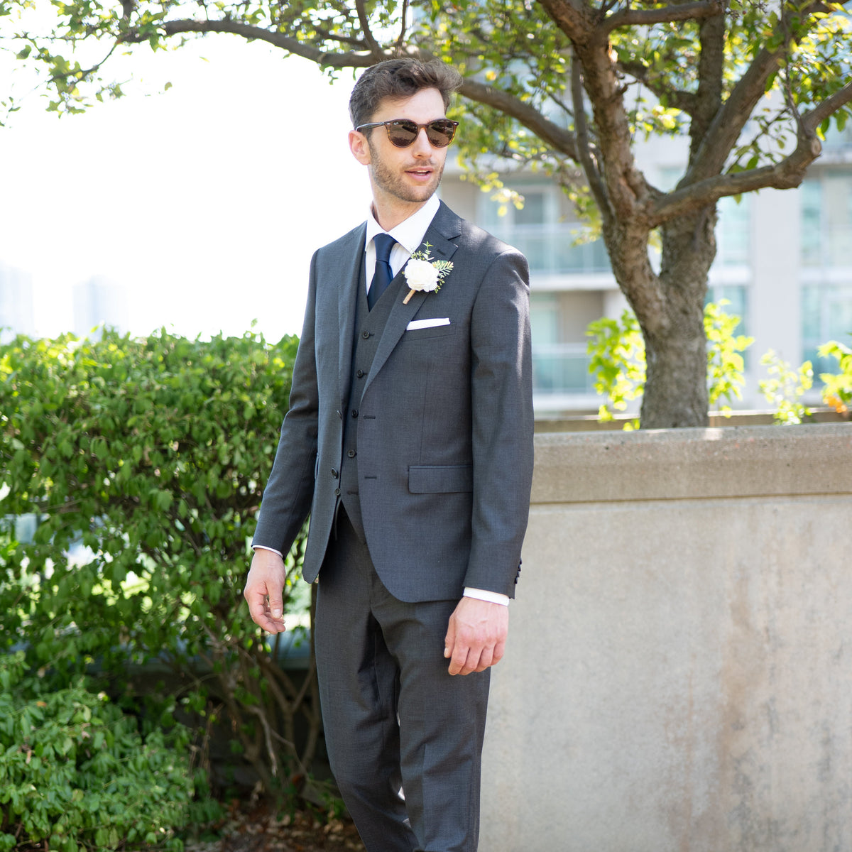 A man wearing a dark gray three-piece suit, white dress shirt, navy tie, and a white rose boutonniere, standing outdoors in front of a concrete wall and greenery.