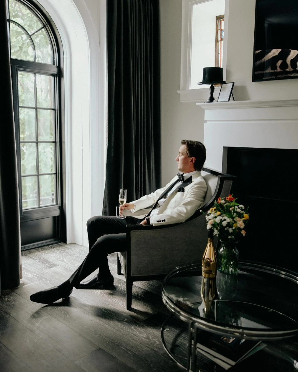 Man in formal attire sitting in a stylish room with a fireplace and large window.