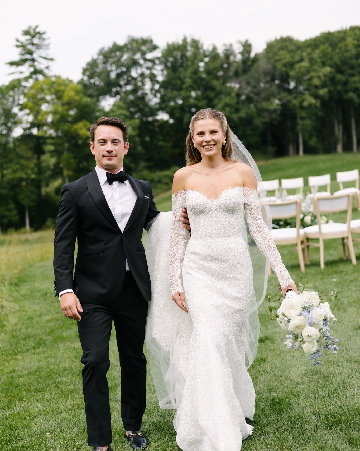 Man in a tuxedo and woman in a white wedding dress standing outdoors with trees and chairs in the background.