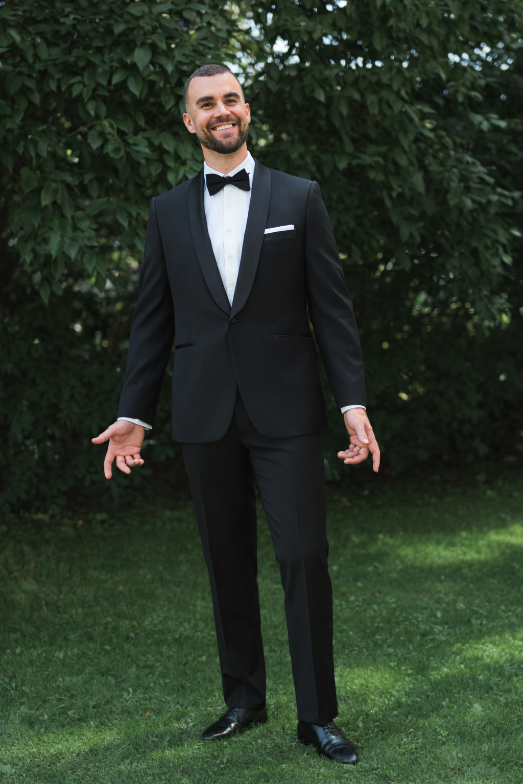 A man wearing a classic black tuxedo with a white dress shirt, black bow tie, and white pocket square, standing outdoors on a green lawn with leafy trees in the background.