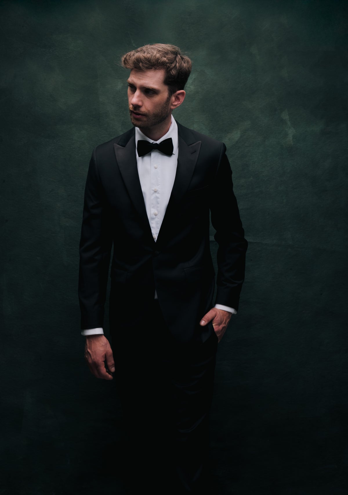 A man wearing a classic black tuxedo with a white dress shirt and black bow tie, standing against a dark textured background.
