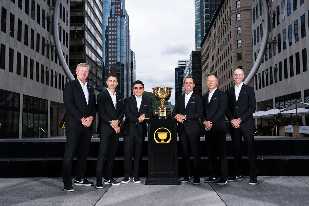 six me in black custom Jack Victor suit stand outdoors in a city setting behind a black pedestal holding a gold trophy cup. Tall buildings and a curved metal sculpture are visible in the background.