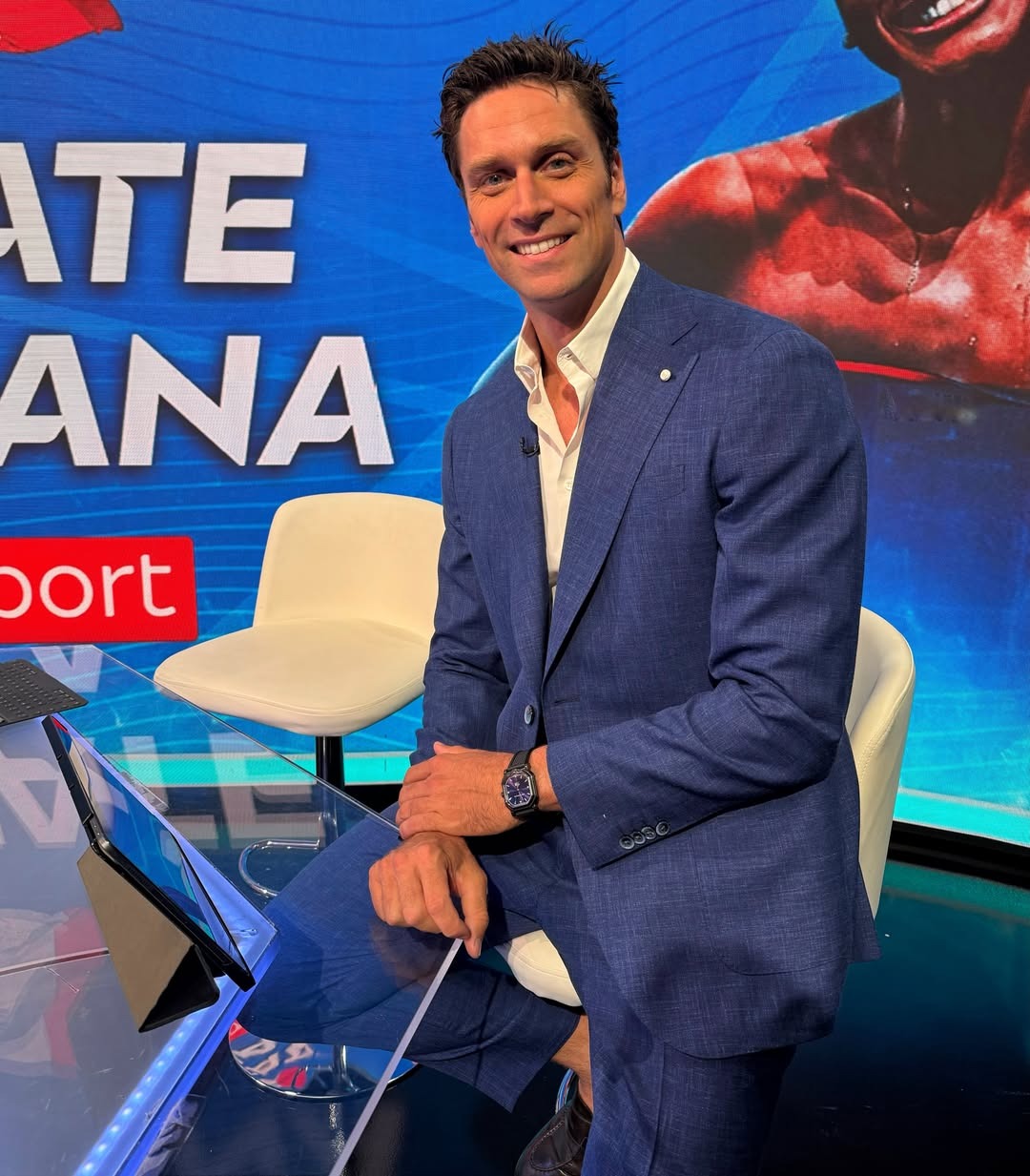 A man in a blue suit and white shirt sits on a white chair in a studio setting, with a blue backdrop featuring large text and a muscular athlete's photo.