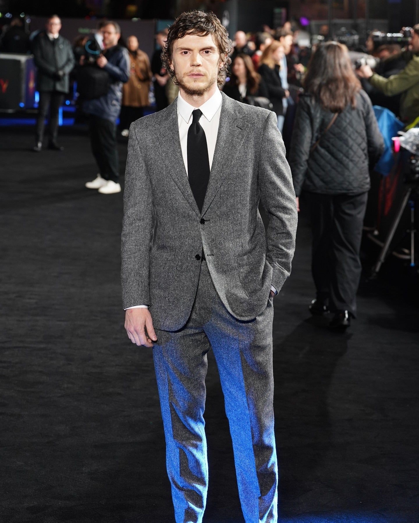 A man wearing a gray textured suit with blue side panels, a white dress shirt, and a black tie, standing on a black carpeted event floor with a blurred crowd in the background.