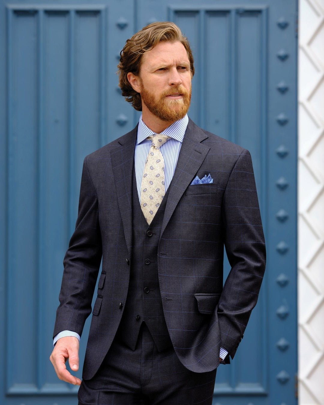 Man wearing a dark suit with a light blue shirt and patterned tie against a blue wall.