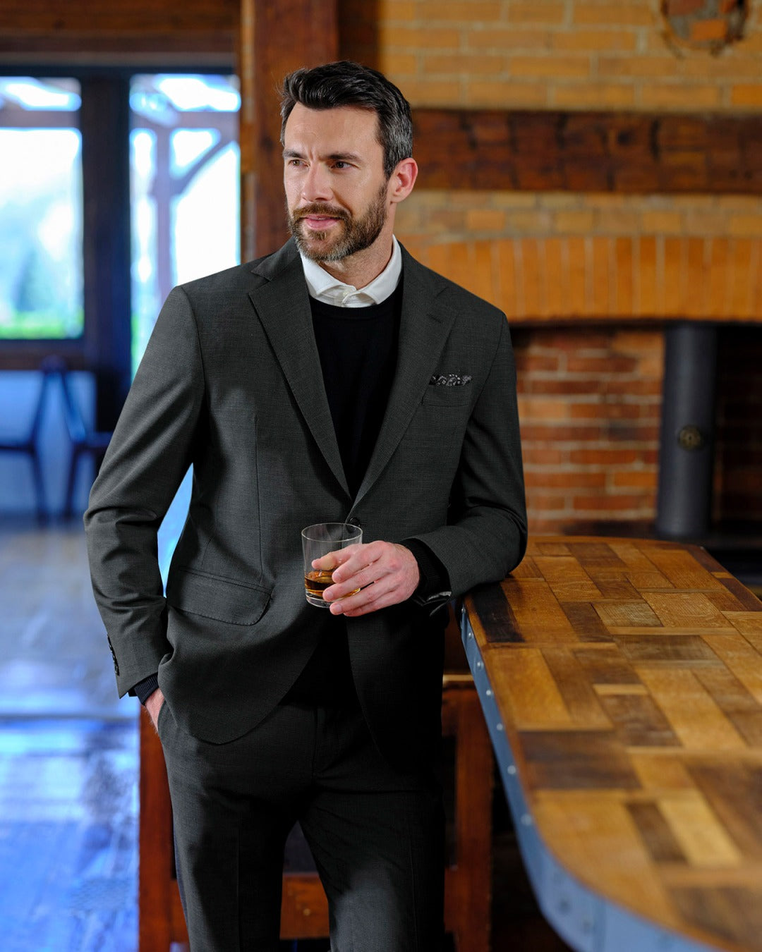 Man in a gray suit holding a drink in a rustic setting