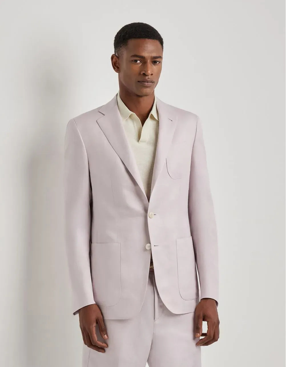 A man wearing a light pink suit jacket and matching trousers over a cream-colored collared shirt, standing against a plain light gray studio background.