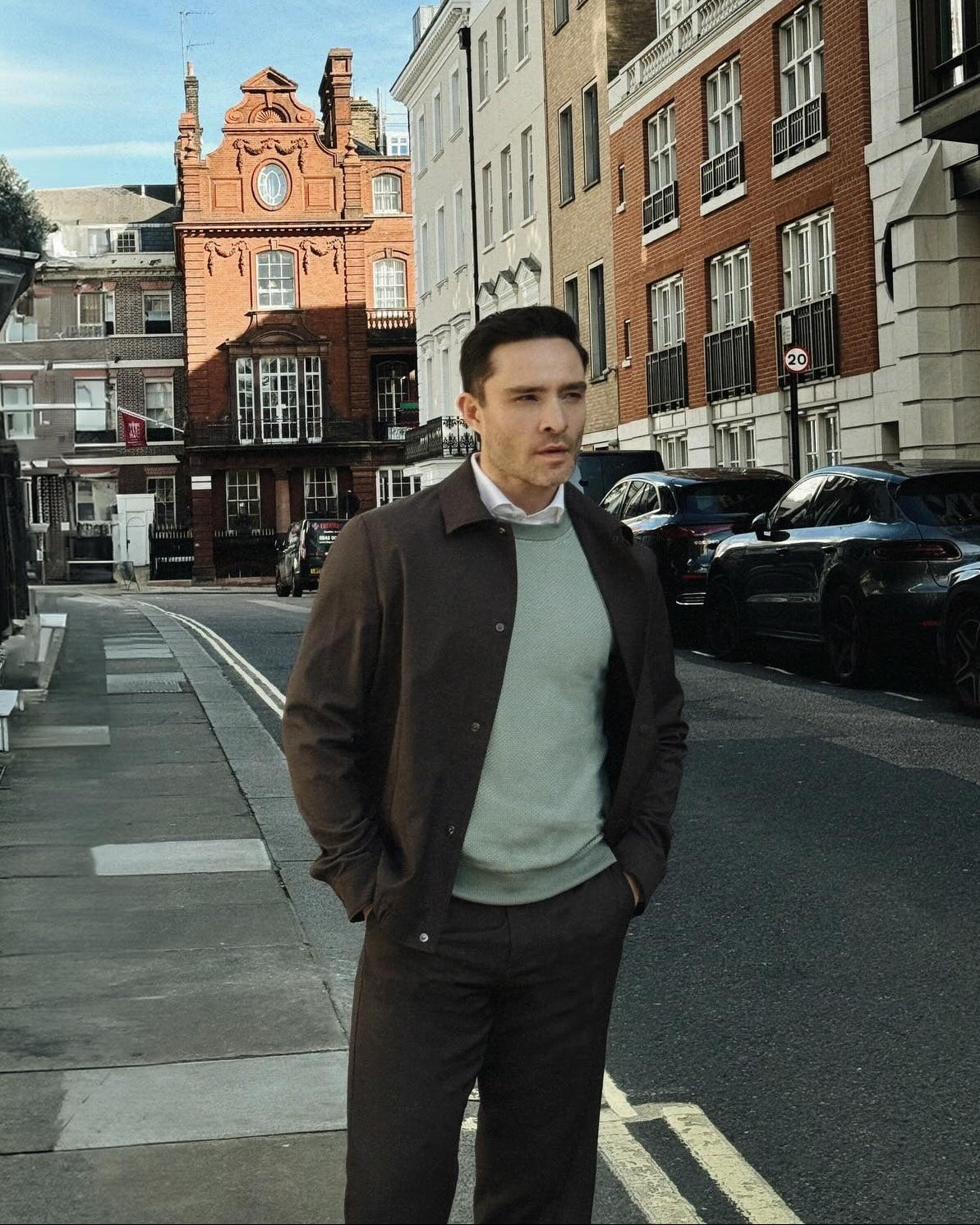 A man wearing a brown jacket over a light green sweater and dark trousers stands on a city street with brick buildings and parked cars in the background.