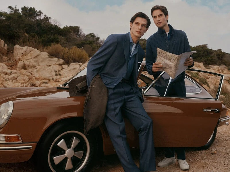 Two men in tailored suits pose beside a brown classic car outdoors on a rocky hillside. One man leans against the car holding a brown bag, while the other stands by the open door holding a map.